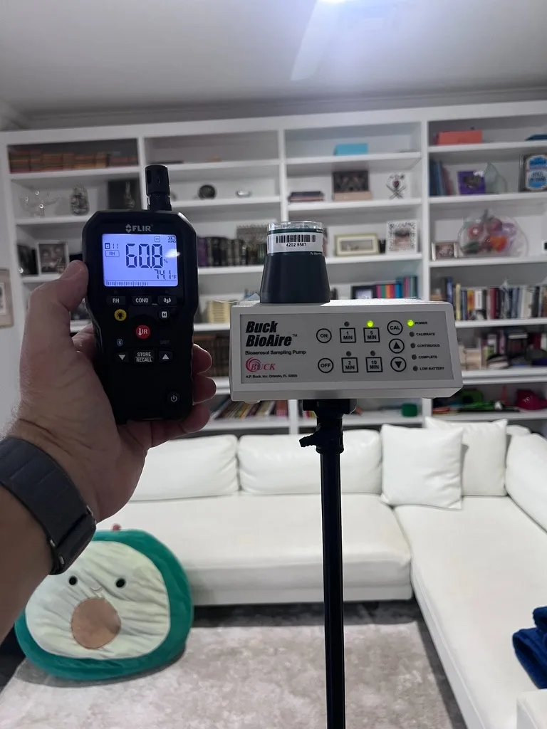 Home Enviro inspector holding a FLIR thermo-hygrometer next to a Buck BioAire air sampling pump on a tripod in a living room