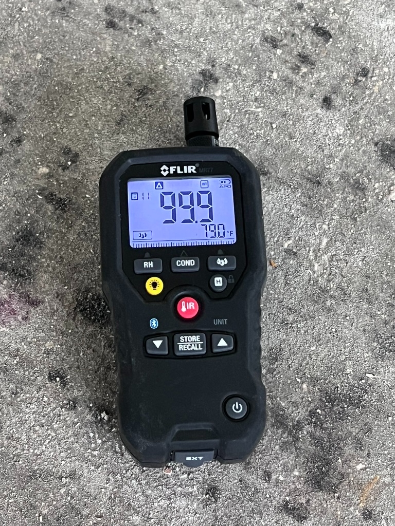 Handheld hygrometer displaying a 99.9 percent relative humidity reading on a concrete garage floor