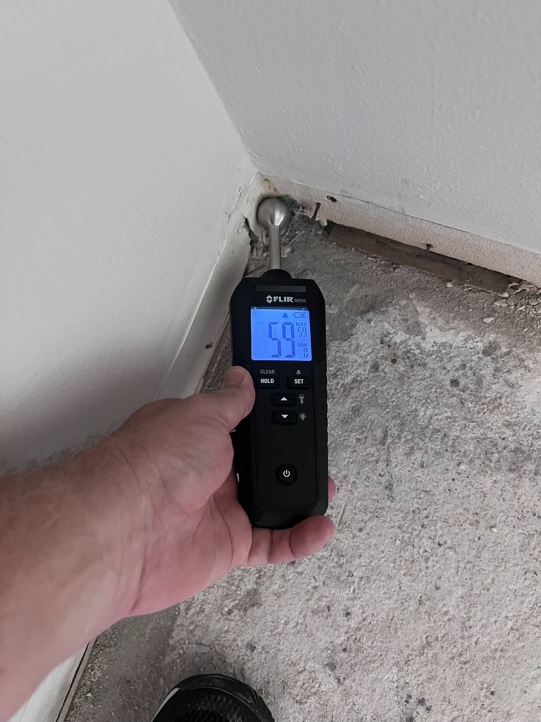 Digital moisture meter reading a high moisture percentage against a wall corner near the slab